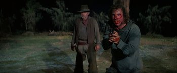 Movie still from “The Cowboys” (1972), directed by Mark Rydell – A man holding a gun and a man with blood on his face; Medium shot, Over the shoulder angle
