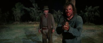 Movie still from “The Cowboys” (1972), directed by Mark Rydell – A man holding a gun while standing next to another man; Medium shot, Over the shoulder angle