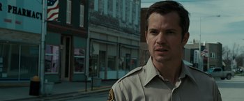 Movie still from “The Crazies” (2010), directed by Breck Eisner – A man in uniform standing in front of some buildings; Close Up shot, Over the shoulder angle