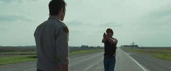 Movie still from “The Crazies” (2010), directed by Breck Eisner – A man holding a gun in front of another man on the side of the road; Wide shot, Over the shoulder angle