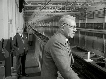 Movie still from “The Dam Busters” (1955), directed by Michael Anderson – A man standing next to another man near a body of water; Medium shot, Low angle