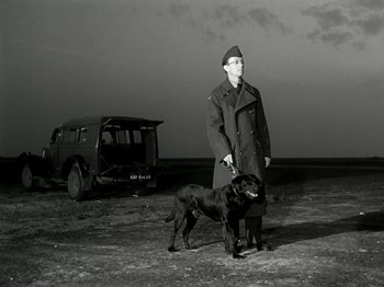 Movie still from “The Dam Busters” (1955), directed by Michael Anderson – A man and a dog standing in front of a truck; Wide shot, Low angle