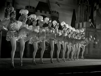 Movie still from “The Dam Busters” (1955), directed by Michael Anderson – A group of women dressed in costumes on a stage; Wide shot, Low angle