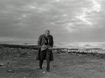 Movie still from “The Dam Busters” (1955), directed by Michael Anderson – A black and white photo of a man standing on the beach; Wide shot, Low angle