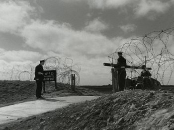Movie still from “The Dam Busters” (1955), directed by Michael Anderson – A black and white photo of two men standing in front of barbed wire; Wide shot, Low angle