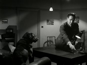 Movie still from “The Dam Busters” (1955), directed by Michael Anderson – A man sitting at a table with a dog; Medium shot, High angle