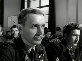 Movie still from “The Dam Busters” (1955), directed by Michael Anderson – Black and white photograph of a man with a mustache in front of a group of other men; Close Up shot, Low angle