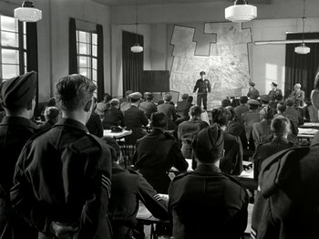 Movie still from “The Dam Busters” (1955), directed by Michael Anderson – A group of people in a room with a man standing in front of them; Wide shot, High angle
