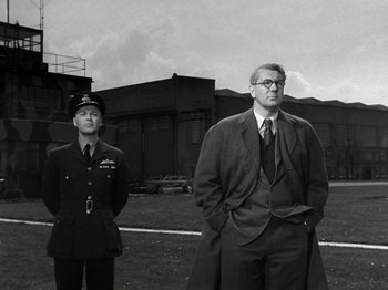 Movie still from “The Dam Busters” (1955), directed by Michael Anderson – A man in a suit stands next to another man in a uniform; Medium shot, Low angle