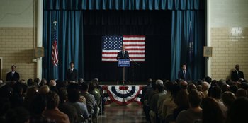 Movie still from “House of Cards” (2013), created by Beau Willimon – A man standing at a podium in front of an american flag; Extreme Wide shot, High angle