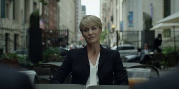 Movie still from “House of Cards” (2013), created by Beau Willimon – A woman sitting at an outdoor table in a city setting; Medium shot, Over the shoulder angle