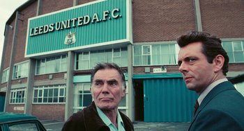 Movie still from “The Damned United” (2009), directed by Tom Hooper – Two men standing in front of a soccer stadium; Medium shot, Over the shoulder angle