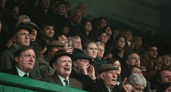 Movie still from “The Damned United” (2009), directed by Tom Hooper – A group of people sitting in the stands at a sporting event; Medium shot, Over the shoulder angle