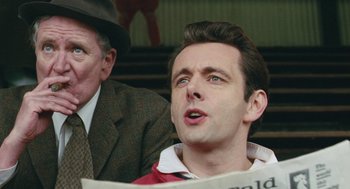 Movie still from “The Damned United” (2009), directed by Tom Hooper – A man sitting next to another man while holding a newspaper; Close Up shot, Over the shoulder angle
