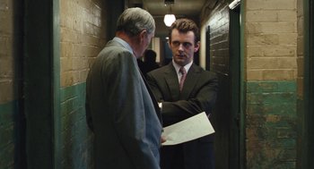Movie still from “The Damned United” (2009), directed by Tom Hooper – A man in a suit and tie standing next to another man in a suit and tie; Medium shot, Over the shoulder angle