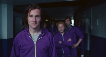 Movie still from “The Damned United” (2009), directed by Tom Hooper – A group of people in purple jackets standing in a room; Medium shot, Over the shoulder angle