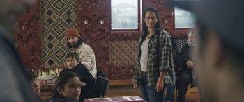 Movie still from “The Dark Horse” (2014), directed by James Napier Robertson – A woman standing in front of a group of people in a room; Medium shot, Over the shoulder angle