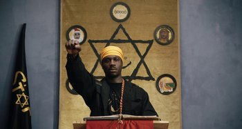 Movie still from “The Day Shall Come” (2019), directed by Christopher Morris – A man in a yellow turban is giving a speech; Medium shot, Low angle
