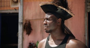 Movie still from “The Day Shall Come” (2019), directed by Christopher Morris – A man wearing a hat with feathers on top of his head; Close Up shot, Low angle