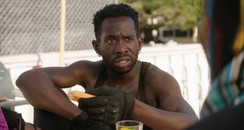 Movie still from “The Day Shall Come” (2019), directed by Christopher Morris – A man sitting on the ground holding a donut; Close Up shot, Over the shoulder angle
