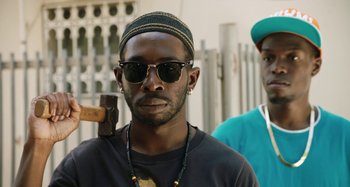 Movie still from “The Day Shall Come” (2019), directed by Christopher Morris – A man wearing sunglasses and holding a hammer; Close Up shot, Low angle