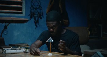 Movie still from “The Day Shall Come” (2019), directed by Christopher Morris – A man sitting at a table making a paper airplane; Medium shot, High angle
