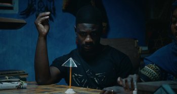 Movie still from “The Day Shall Come” (2019), directed by Christopher Morris – A man sitting at a table holding a paper umbrella; Medium shot, Low angle