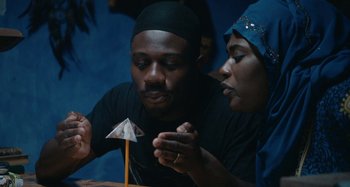 Movie still from “The Day Shall Come” (2019), directed by Christopher Morris – A man and a woman looking at an object on top of a table; Medium shot, High angle