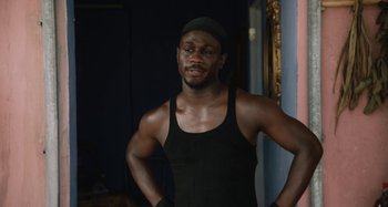Movie still from “The Day Shall Come” (2019), directed by Christopher Morris – A man in a black tank top is posing for a picture; Medium shot, Over the shoulder angle