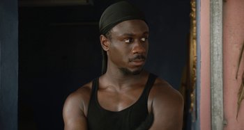 Movie still from “The Day Shall Come” (2019), directed by Christopher Morris – A man in a black tank top wearing a black hat; Close Up shot, Over the shoulder angle