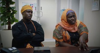 Movie still from “The Day Shall Come” (2019), directed by Christopher Morris – A man and a woman sitting in front of a wooden table; Medium shot, Over the shoulder angle