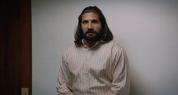 Movie still from “The Day Shall Come” (2019), directed by Christopher Morris – A man with long hair wearing a striped shirt; Medium shot, Over the shoulder angle