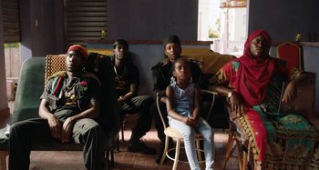 Movie still from “The Day Shall Come” (2019), directed by Christopher Morris – A group of people sitting in a room; Medium shot, Over the shoulder angle