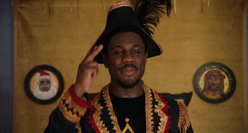 Movie still from “The Day Shall Come” (2019), directed by Christopher Morris – A man wearing a black and gold jacket and a black hat; Close Up shot, Low angle