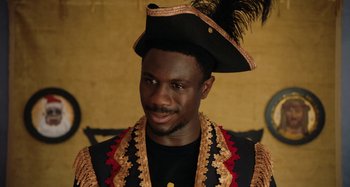 Movie still from “The Day Shall Come” (2019), directed by Christopher Morris – A man wearing a black and gold outfit with a black hat; Close Up shot, Low angle