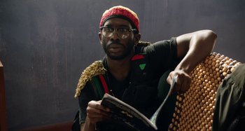Movie still from “The Day Shall Come” (2019), directed by Christopher Morris – A man in a red hat and glasses is reading a magazine; Close Up shot, Low angle