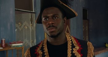 Movie still from “The Day Shall Come” (2019), directed by Christopher Morris – A man wearing a black and gold outfit and wearing a black hat; Close Up shot, Low angle