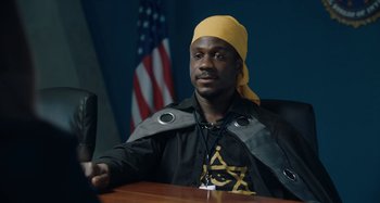 Movie still from “The Day Shall Come” (2019), directed by Christopher Morris – A man sitting in front of an american flag; Medium shot, Over the shoulder angle