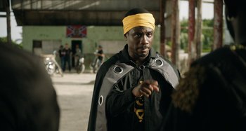 Movie still from “The Day Shall Come” (2019), directed by Christopher Morris – A man wearing a yellow bandanna standing in a parking lot; Medium shot, Over the shoulder angle