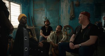 Movie still from “The Day Shall Come” (2019), directed by Christopher Morris – A group of men sitting in a room; Medium shot, Low angle