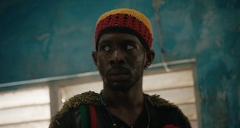 Movie still from “The Day Shall Come” (2019), directed by Christopher Morris – A man wearing a hat with a yellow , red , and green hat; Close Up shot, Low angle