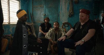 Movie still from “The Day Shall Come” (2019), directed by Christopher Morris – A group of men sitting in a room together; Medium shot, Over the shoulder angle