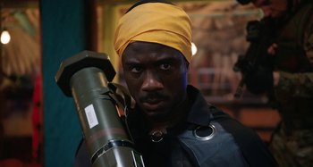 Movie still from “The Day Shall Come” (2019), directed by Christopher Morris – A man with a yellow head band holding a large object; Close Up shot, Over the shoulder angle