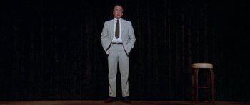 Movie still from “The Day of the Dolphin” (1973), directed by Mike Nichols – A man standing on a stage wearing a suit and tie; Wide shot, Low angle