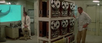 Movie still from “The Day of the Dolphin” (1973), directed by Mike Nichols – A man standing in front of a row of old tape recorders; Wide shot, High angle