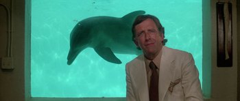 Movie still from “The Day of the Dolphin” (1973), directed by Mike Nichols – A man standing in front of an aquarium with a dolphin swimming in the background; Medium shot, Low angle
