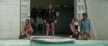 Movie still from “The Day of the Dolphin” (1973), directed by Mike Nichols – A man standing on top of a surfboard in the water; Wide shot, Low angle