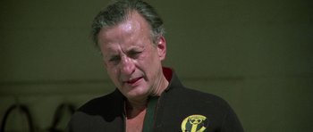 Movie still from “The Day of the Dolphin” (1973), directed by Mike Nichols – An older man wearing a black jacket with a yellow and black logo on it's chest; Close Up shot, Low angle