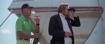 Movie still from “The Day of the Dolphin” (1973), directed by Mike Nichols – A man taking a picture of another man holding a cigar; Medium shot, Over the shoulder angle