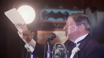 Movie still from “The Day of the Locust” (1975), directed by John Schlesinger – A man in a suit and tie holding a microphone; Close Up shot, Over the shoulder angle